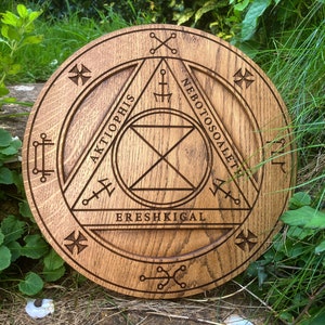 Puede incluir: Una placa circular de madera con un diseño tallado de un triángulo con las palabras "AKTIOPHIS", "NEBOTOSOALETH" y "ERESHIKIGAL" inscritas alrededor.