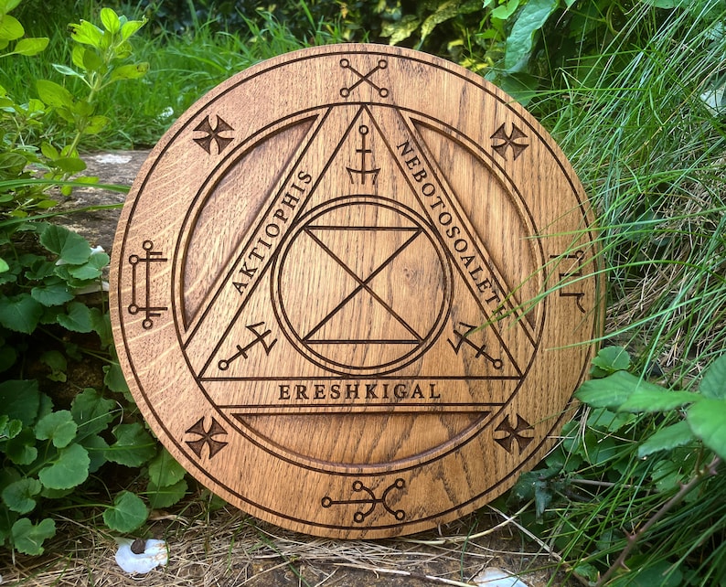 Op de afbeelding: Een ronde houten plaquette met een gesneden ontwerp van een driehoek met de woorden "AKTIOPHIS", "NEBOTOSOALETH" en "ERESHIKIGAL" eromheen gegraveerd.