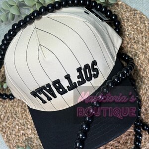 May include: A cream-colored softball cap with black pinstripes and a black brim. The word "SOFTBALL" is embroidered in black on the front. A black beaded necklace is draped around the cap.