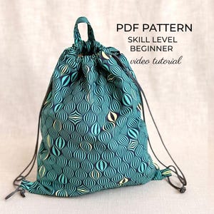 Puede incluir: Una mochila de cordón con un patrón geométrico en azul cerceta, azul oscuro y crema. La bolsa tiene un asa superior y correas de cordón marrón oscuro. El texto en la imagen dice: PDF PATTERN, SKILL LEVEL BEGINNER, video tutorial.