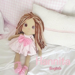 May include: A handmade doll with brown and blonde yarn hair, large brown eyes, and a pink dress with a bow. The doll is wearing pink ballet flats. The word "Hannita" and the word "English" are written in pink.