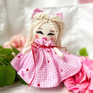 May include: A handmade fabric doll with blonde curly hair and a pink gingham dress adorned with small red hearts. The doll has rosy cheeks, closed eyes, and a red ribbon tied around its neck. A pink bow sits atop the doll's head.