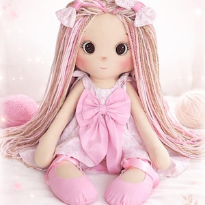 May include: A handmade rag doll with long, braided hair in shades of pink and tan. The doll wears a pink dress with a large bow and matching pink shoes. Two small bows adorn the doll's hair. The doll is sitting on a white surface.
