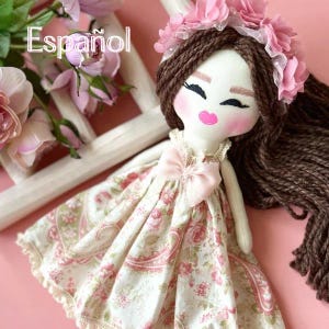 以下が含まれることがあります： 茶色の糸の髪とピンクの花の冠が付いた手作りの布製人形。人形はピンクのバラ模様のドレスとピンクのリボンを着ています。「Español」という言葉が見えます。