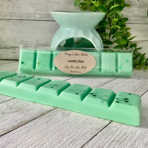 May include: Two green wax melt bars with a light green wax warmer in the background. The wax melts are labeled "Laundry Gain" and are speckled with darker green flecks. The wax warmer is a light blue color.