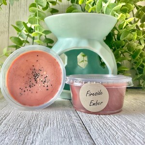 May include: Two containers of wax melts with a light pink color and glitter. One container is open, and the other has a label that reads "Fireside Ember." A light green wax warmer is in the background.
