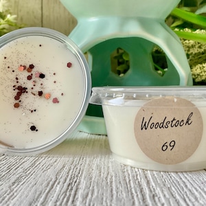May include: Two small, clear plastic containers filled with off-white wax melts. One is open, sprinkled with glitter. The other has a label that reads "Woodstock 69". A light green ceramic oil warmer is in the background.