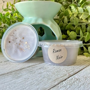 May include: A light purple wax melt with gold glitter accents in a clear plastic container labeled Roman Noir, positioned next to a light green ceramic wax warmer. The wax melt has a smooth surface with visible gold flecks scattered on top.