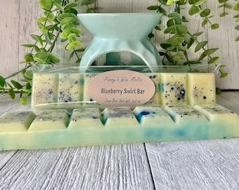 May include: A light blue and yellow wax melt bar with blue speckles, labeled "Blueberry Swirl Bar." The bar is in a clear plastic package, with a light blue ceramic oil warmer in the background. The bar weighs 3.2 oz.