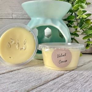May include: A light yellow wax melt in a clear plastic container with the text "Velvet Cream" on a round label. A light blue ceramic wax warmer is in the background. The wax melt has a creamy appearance and is surrounded by greenery.