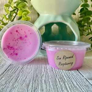 May include: Two small, clear plastic containers filled with pink wax melts. One is open, revealing a speckled surface. The other has a lid and a label that reads "Sun Ripened Raspberry." A light green wax warmer is in the background.