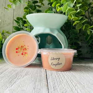 May include: Two small, clear plastic containers of peach-colored wax melts with a light dusting of red and yellow sprinkles. One container has a label that reads "Library Fireplace." A light blue ceramic wax warmer is in the background.