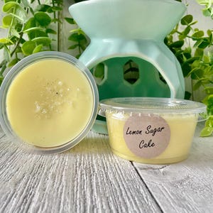 May include: Two small containers of wax melts, one with a yellow wax melt and the other with a yellow wax melt and a label that says "Lemon Sugar Cake".