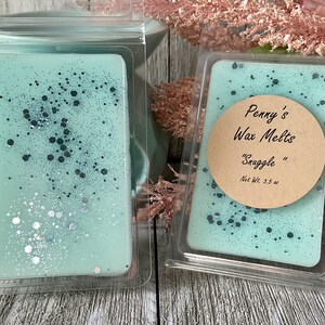 May include: Two wax melt bars in clear plastic packaging. The wax is a light teal color, speckled with dark blue and silver glitter. One bar has a round label that reads "Penny's Wax Melts" and "Snuggle". Each bar weighs 3.5 oz.