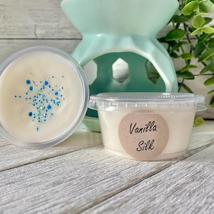 May include: Two small, clear plastic containers of wax melts. One is open, showing a creamy white wax with blue glitter. The other is closed, with a label that reads "Vanilla Silk". A light blue wax warmer is in the background.
