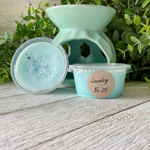 May include: A light blue wax melt with glitter, next to a matching teal ceramic oil warmer. A small container of wax melt is labeled "Laundry No 26" on a round sticker. The items are on a white wooden surface.