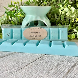 May include: A light blue wax melt bar with a segmented rectangular shape and speckled texture. An oval label reads Penny's Wax Melts, Laundry No. 26, Soap Bar Wax Melt, Net Wt. 2.8 oz. The wax melt is positioned in front of a light blue wax warmer.