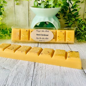 May include: A yellow wax melt bar with a cornbread scent, featuring a segmented design. The bar is labeled "Nana's Cornbread Soap Bar Wax Melt" and is packaged in clear plastic. The background includes greenery and a light blue wax warmer.
