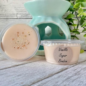 May include: Two small, clear plastic containers of Vanilla Sugar Bonbon wax melts. One is open, revealing a light peach-colored wax with gold glitter. A light blue ceramic wax warmer is in the background.