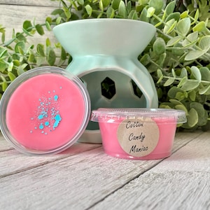 May include: A light blue ceramic wax warmer with a round opening, next to two containers of pink wax melts. One melt has blue sprinkles, and the other has a label that reads "Cotton Candy Maniac."