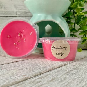 May include: Two containers of pink wax melts with gold flakes, one open and one with a label that reads "Strawberry Candy." A light green essential oil diffuser is in the background, along with green foliage. The melts are on a white wooden surface.