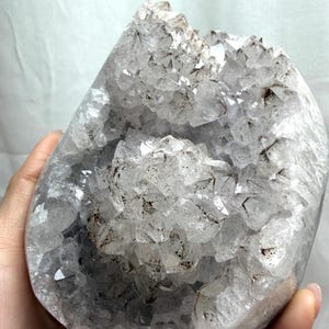May include: A large, rounded quartz geode featuring a cluster of translucent, multifaceted crystals. The interior displays clear, pointed crystal formations with dark, speckled inclusions scattered throughout the structure, creating a textured appearance.