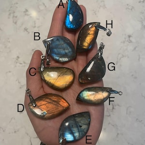 以下が含まれることがあります： 銀色のベイルが付いた8つのラブラドライトペンダントのコレクション。石は、青、オレンジ、緑など、さまざまな色を示し、虹色のフラッシュがあります。各ペンダントはユニークな形とパターンを持ち、天然石のバリエーションを示しています。寸法はcmです。