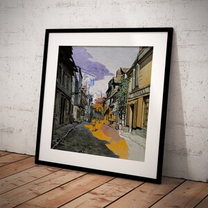 May include: Framed art print of a European street scene. The artwork depicts a narrow street with buildings on either side, a few figures, and a bright sky. The print is in a black frame and leans against a white brick wall.