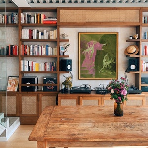 K&ouml;nnte beinhalten: Ein Holztisch mit rustikaler Oberfl&auml;che im Vordergrund. Dahinter ein Holzschrank mit Regalen, gef&uuml;llt mit B&uuml;chern und Dekorationsgegenst&auml;nden. Ein gerahmtes Kunstwerk in Gr&uuml;n- und Rosat&ouml;nen h&auml;ngt &uuml;ber dem Schrank. Eine Vase mit Blumen steht auf dem Tisch.
