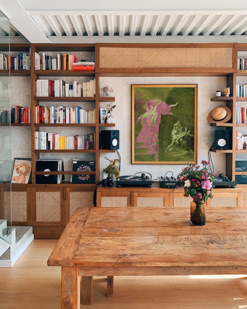 K&ouml;nnte beinhalten: Ein Holztisch mit rustikaler Oberfl&auml;che im Vordergrund. Dahinter ein Holzschrank mit Regalen, gef&uuml;llt mit B&uuml;chern und Dekorationsgegenst&auml;nden. Ein gerahmtes Kunstwerk in Gr&uuml;n- und Rosat&ouml;nen h&auml;ngt &uuml;ber dem Schrank. Eine Vase mit Blumen steht auf dem Tisch.