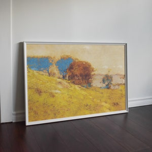 May include: A framed landscape painting featuring a hillside with trees. The artwork displays a palette of yellow, blue, and brown hues, with a silver frame. The painting leans against a white wall and dark wood floor.
