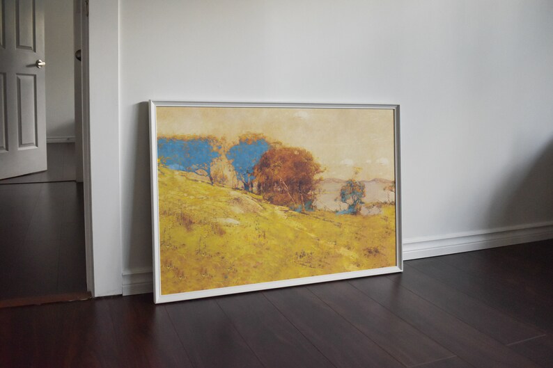 May include: A framed landscape painting featuring a hillside with trees. The artwork displays a palette of yellow, blue, and brown hues, with a silver frame. The painting leans against a white wall and dark wood floor.