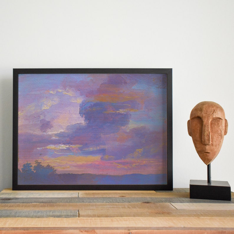 May include: A framed landscape painting with a black frame depicts a sunset with purple, blue, and gold hues. A wooden sculpture of a face sits on a black stand to the right, placed on a wooden surface.