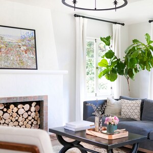 K&ouml;nnte beinhalten: Wohnzimmer mit blauem Ecksofa, schwarzem Couchtisch und Kamin mit gestapeltem Brennholz. Ein gerahmtes Blumenbild h&auml;ngt &uuml;ber dem Kamin. Eine gro&szlig;e Topfpflanze und wei&szlig;e Vorh&auml;nge sind ebenfalls zu sehen.