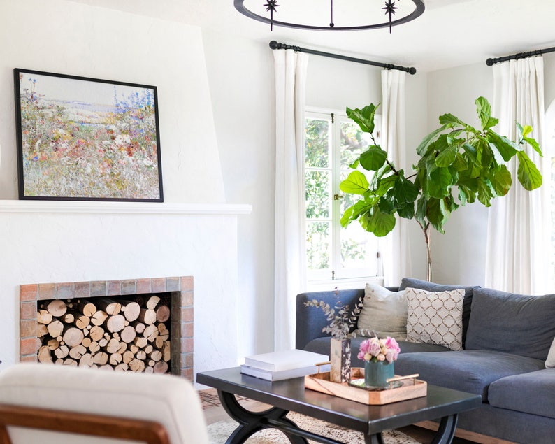 K&ouml;nnte beinhalten: Wohnzimmer mit blauem Ecksofa, schwarzem Couchtisch und Kamin mit gestapeltem Brennholz. Ein gerahmtes Blumenbild h&auml;ngt &uuml;ber dem Kamin. Eine gro&szlig;e Topfpflanze und wei&szlig;e Vorh&auml;nge sind ebenfalls zu sehen.
