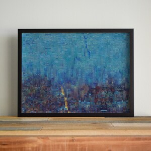May include: Framed abstract art print depicting a cityscape with a blue and teal color palette. The artwork features a textured, pixelated effect, with hints of gold and brown. The black frame sits on a wooden surface.