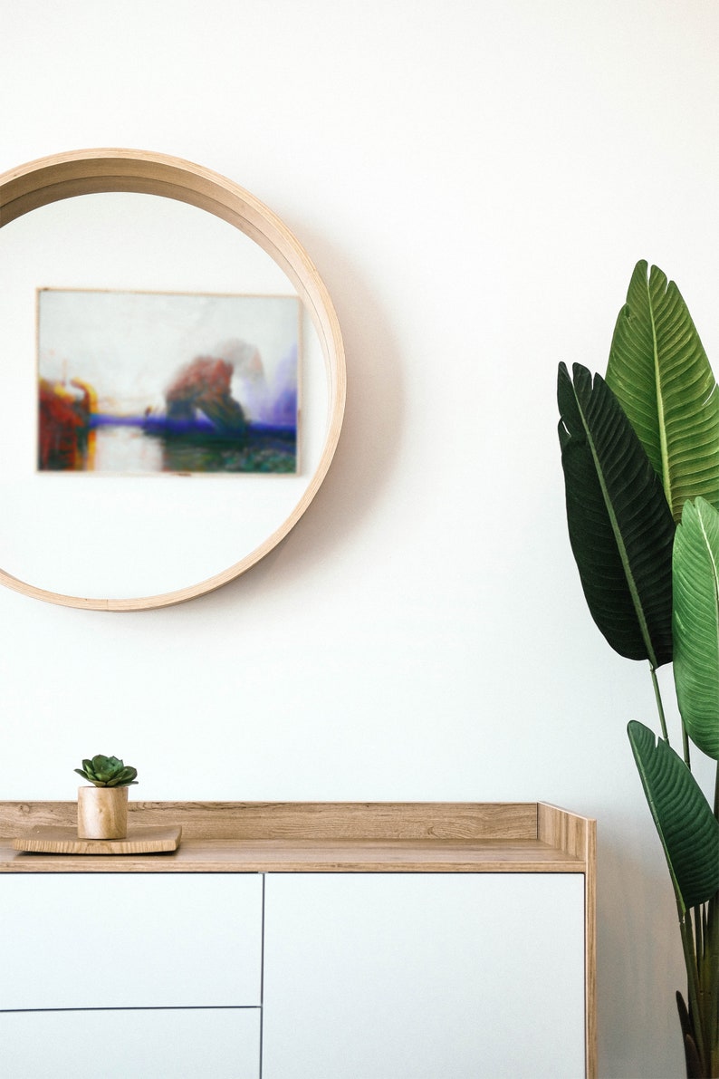 May include: A contemporary interior scene with a circular wooden mirror displaying a blurred painting, a white cabinet with wooden detailing, a small succulent plant, and large green leaves. The design creates a minimalist and natural aesthetic.
