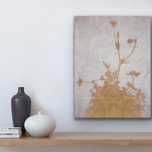 May include: A gold-toned floral print on a canvas, featuring delicate plant silhouettes against a textured, light gray background. The artwork is displayed on a wooden shelf, with a black vase, a white vase, and a stack of books.