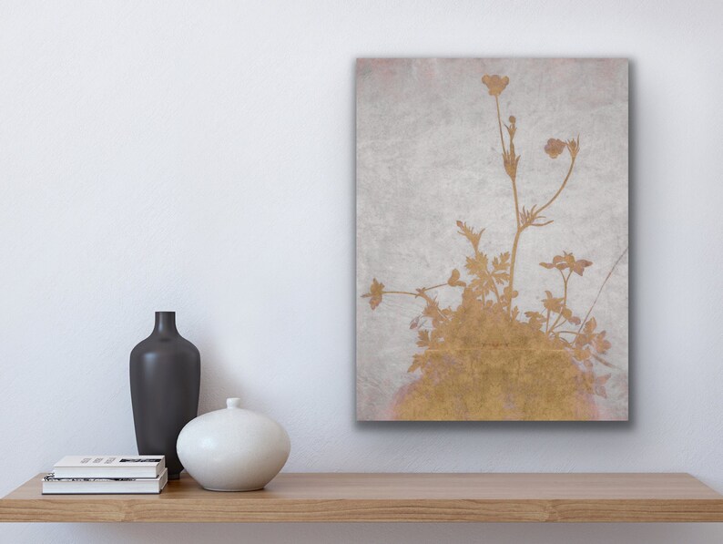 May include: A gold-toned floral print on a canvas, featuring delicate plant silhouettes against a textured, light gray background. The artwork is displayed on a wooden shelf, with a black vase, a white vase, and a stack of books.