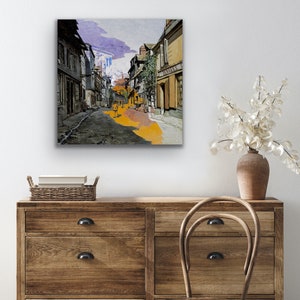 May include: An art print of a European street scene. The painting depicts buildings lining a cobblestone street, with a vibrant yellow and orange light illuminating the scene. The sky is a mix of purple and white, and two figures are walking down the street.