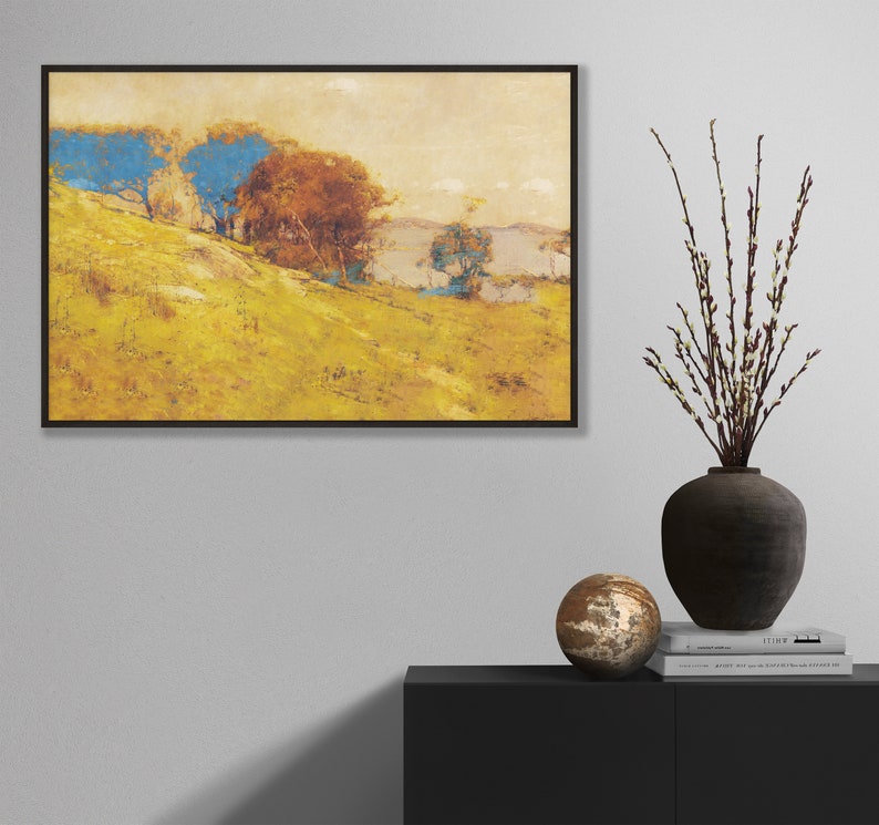May include: An art print in a black frame depicts a landscape with a yellow hillside, blue and brown trees, and a body of water in the background. A dark vase with branches and a decorative sphere sit on a black cabinet.