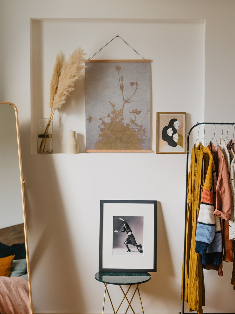 May include: A room interior featuring framed art and decorative items. A large framed print of a dancer is on a small table. Above, a botanical print hangs, and a smaller artwork depicts three figures. A clothing rack with various garments is on the right.