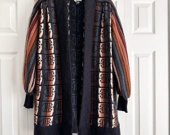 May include: A long, open-front cardigan in a geometric pattern. The sweater features a black base with a repeating pattern in shades of brown, blue, and white. The sleeves have a striped design in similar colors. The cardigan is hanging on a black hanger.