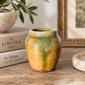 May include: A small, rounded ceramic vase featuring a glossy glaze with a blend of earthy mustard yellow and sage green tones. The surface displays a fine crackle pattern throughout, with a wide, open mouth and a smooth, tapered base.