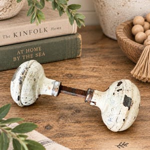 May include: A vintage-style door knob set featuring two round, off-white ceramic or metal knobs with a distressed, chipped paint finish revealing dark metal underneath. The two knobs are connected by a central, square-shaped dark metal spindle rod.