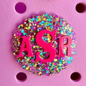 May include: A pink, scalloped-edge charm with the letters "ASR" in a vibrant pink hue. The charm is filled with colorful glitter and sits on a light pink surface with circular cutouts. The letters are raised, creating a 3D effect.