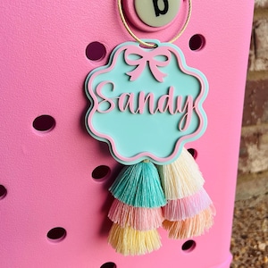 Puede incluir: Una etiqueta de nombre personalizada de color azul claro y rosa con el nombre "Sandy" escrito en letras cursivas rosas. La etiqueta tiene un lazo rosa y está unida a un borla multicolor.