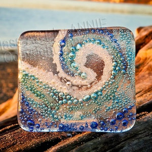May include: A square glass coaster featuring a swirling spiral pattern of numerous small, raised bubbles in shades of deep blue, turquoise, and white. The clear glass surface displays a textured, multi-layered design with varying bubble sizes.