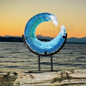 May include: A blue glass wave sculpture on a black metal stand. The sculpture is shaped like a circle with a gap in the middle, creating a wave-like effect. The sculpture is set against a backdrop of a body of water and a mountain range in the distance.