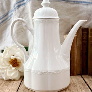 Könnte beinhalten: Eine weiße Porzellan-Kaffeekanne mit Deckel und gebogenem Griff. Die Kanne hat einen langen Ausguss und dekorative Details am Boden. Eine weiße Blume und ein Stapel Bücher befinden sich im Hintergrund.
